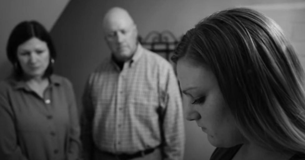 Video still or concerned parents looking toward their daughter.